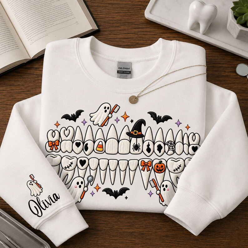 May include: A white crewneck sweatshirt featuring a graphic of human teeth decorated with Halloween icons like ghosts, bats, a pumpkin, and a witch hat. The sleeve displays a ghost holding a toothbrush with the name Olivia written in black script.
