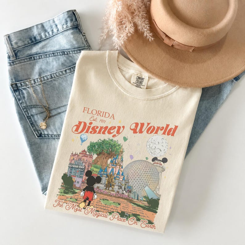 May include: Cream-colored t-shirt with a Disney World graphic, jeans, a gold necklace, and a tan hat. The shirt features the words "Florida Disney World" and a colorful illustration of the theme park. The jeans are light blue.