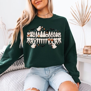 May include: A dark green long-sleeve sweatshirt featuring a central graphic of a set of teeth. The teeth are decorated with small Halloween-themed icons including ghosts, a witch hat, a pumpkin, candy corn, bows, hearts, and a spider web.