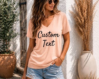 May include: A peach-colored V-neck t-shirt with the words "Custom Text" in black script. The shirt is paired with denim shorts. The image is taken outdoors, with a neutral background and decorative plants.
