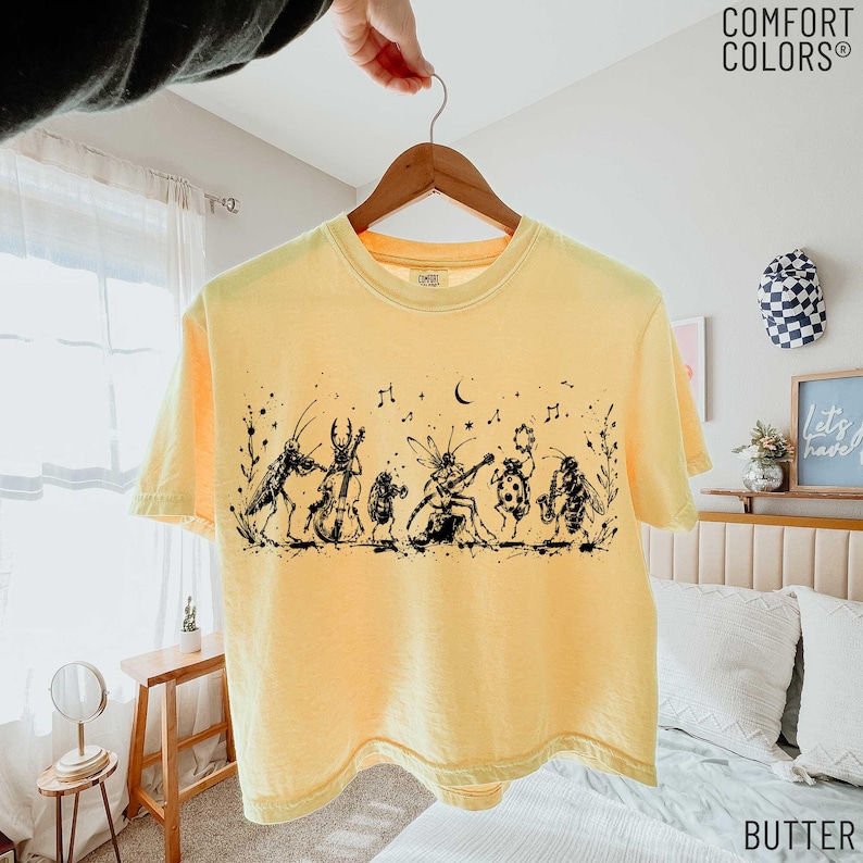 May include: A pale yellow short-sleeve t-shirt featuring a black ink-style graphic of various insects playing musical instruments, including a cricket, beetle, and bee, with musical notes and stars scattered above the figures.