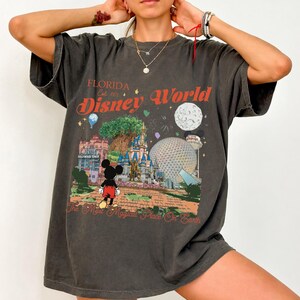 May include: Dark gray t-shirt featuring a colorful Disney World graphic. The design includes the text "Florida Est. 1971 Disney World" and a depiction of Mickey Mouse, a castle, and other park landmarks. The shirt has a vintage, worn look.