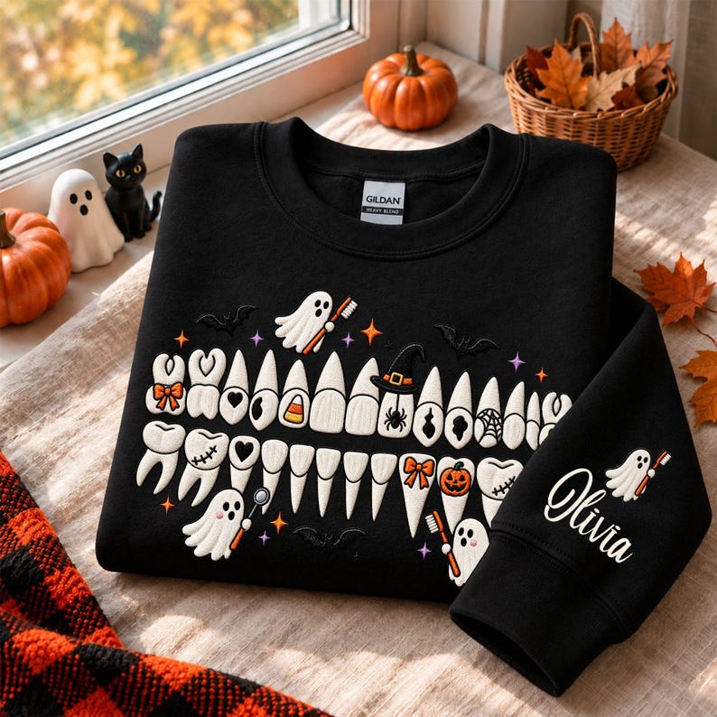May include: A black crewneck sweatshirt featuring an embroidered graphic of teeth decorated with Halloween motifs like ghosts, pumpkins, bats, and spiders. The sleeve is embroidered with the name Olivia and a ghost holding a toothbrush.