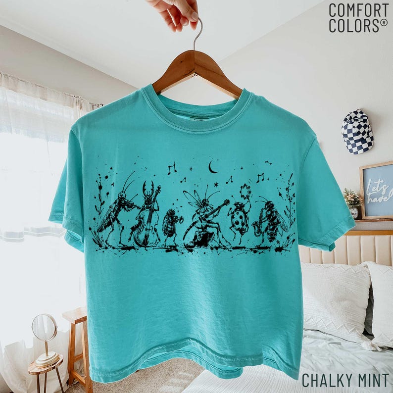 May include: A turquoise t-shirt featuring a black ink-style graphic of various insects playing musical instruments, including a cricket, beetle, and ladybug. The design includes musical notes and floral accents. The shirt has a crew neckline.