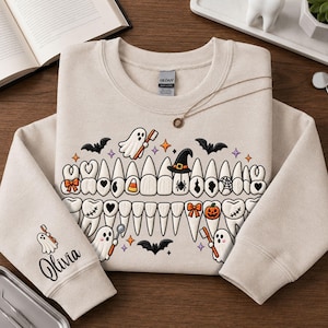 May include: A beige crewneck sweatshirt featuring a graphic of human teeth decorated with Halloween icons like ghosts, bats, pumpkins, and candy corn. The sleeve displays a ghost holding a toothbrush and the name Olivia in black cursive script.