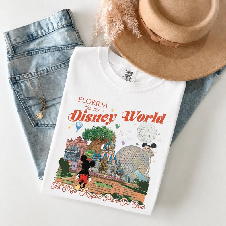 May include: White t-shirt with a colorful graphic of Disney World in Florida. The design includes Mickey Mouse, the Hollywood Tower Hotel, Spaceship Earth, and the text "Florida Disney World" in red and orange. The shirt is laid on top of blue jeans and a tan hat.