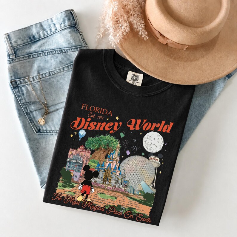May include: Black crew neck t-shirt with a colorful Disney World graphic. The graphic includes Mickey Mouse, a castle, and the text "Florida Disney World Est. 1971". The shirt is paired with blue jeans and a tan hat.