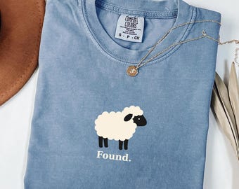 Puede incluir: Una camiseta color azul jean con cuello redondo y etiqueta de la marca Comfort Colors. La prenda presenta un gráfico de una oveja blanca con cara y patas negras, situado sobre el texto blanco Found. Un collar de cadena dorada fina descansa encima.