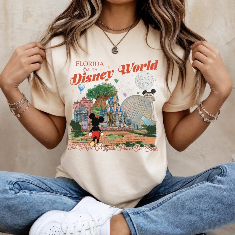 May include: Cream-colored t-shirt featuring a colorful graphic of Disney World in Florida. The design includes the text "Florida Est. 1971 Disney World" and "The Most Magical Place On Earth" with Mickey Mouse in the center.