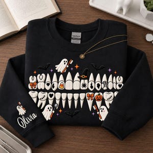 May include: A black long-sleeve sweatshirt featuring a graphic of white teeth decorated with Halloween icons like ghosts, pumpkins, bats, and candy corn. The name Olivia is printed in white script on the left sleeve with a small ghost graphic.