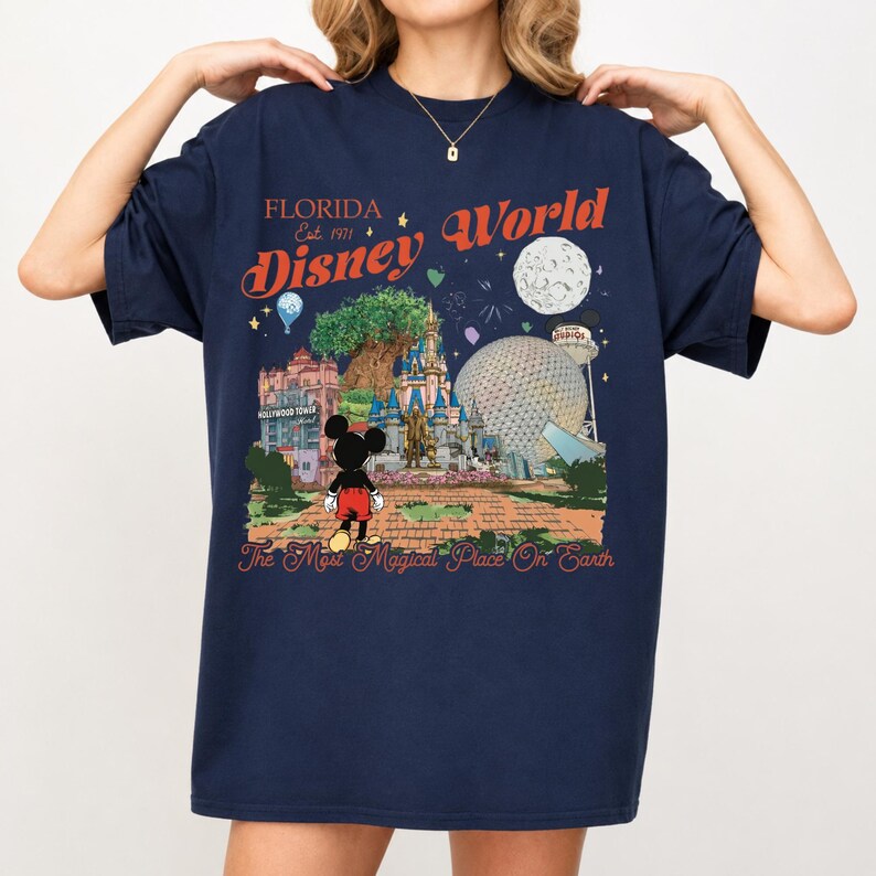May include: Navy blue t-shirt featuring a colorful Disney World graphic. The design includes the text "Florida Disney World Est. 1971" and an illustration of Mickey Mouse, theme park landmarks, and the phrase "The Most Magical Place On Earth."