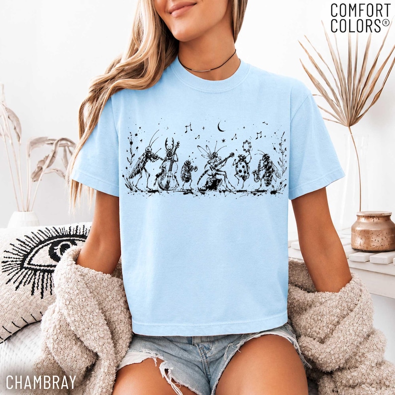 May include: A light blue t-shirt featuring a black ink-style graphic of various insects playing musical instruments, including a cricket, beetle, and bee. The design includes musical notes, a crescent moon, and stars. The text COMFORT COLORS is visible.
