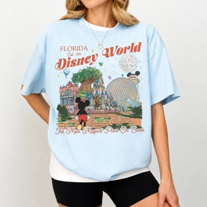 May include: Light blue t-shirt featuring a colorful graphic of Disney World landmarks, including Cinderella's castle and Spaceship Earth. The shirt has the text "Florida Disney World" and "The Most Magical Place On Earth" in a vintage style.