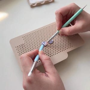 Könnte beinhalten: Ein beiges, rechteckiges Bastelbrett mit einem Raster aus kleinen Löchern, auf dem zwei kleine, gemusterte, kreisförmige Teile mit zwei hellblauen und mintgrünen Präzisionswerkzeugen mit weißen Silikonspitzen bearbeitet werden.