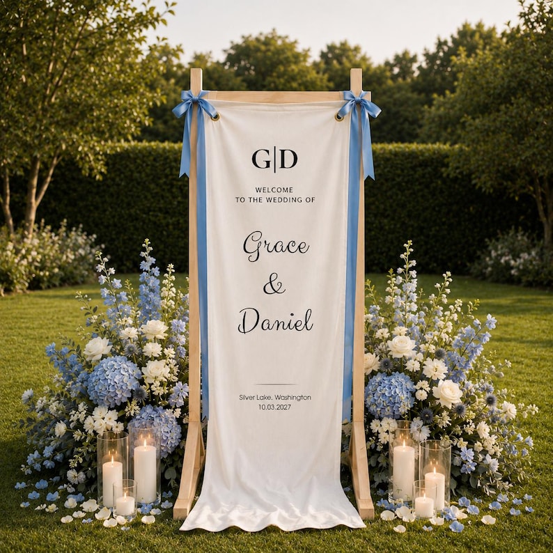 K&ouml;nnte beinhalten: Ein h&ouml;lzernes Hochzeitsschild mit einem wei&szlig;en Stoffbanner. Auf dem Banner steht "G|D WILLKOMMEN ZUR HOCHZEIT VON Grace & Daniel" mit Ort und Datum darunter. Blaue B&auml;nder schm&uuml;cken die oberen Ecken. Das Schild ist von blauen und wei&szlig;en Blumen und Kerzen umgeben.