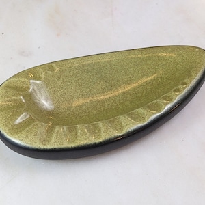 May include: A green ceramic ashtray with a black rim. The ashtray is shaped like a teardrop and has a textured surface.