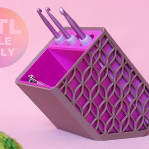 May include: A purple and magenta 3D printed organiser with a lattice side pattern, holding three purple crochet hooks and a small metal clip. The organiser features a slanted top opening and a circular badge stating STL FILE ONLY.