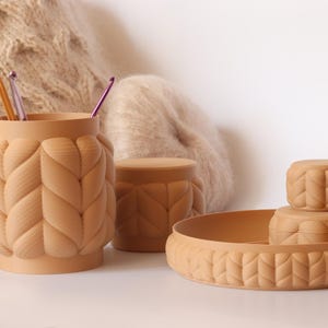 Puede incluir: Un conjunto de recipientes de cerámica beige con un diseño texturizado y tejido. El conjunto incluye un jarrón cilíndrico con agujas de tejer y ganchillos, un tarro pequeño con tapa y una bandeja escalonada. Los artículos están sobre una superficie blanca.