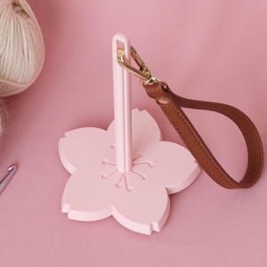 以下が含まれることがあります： ピンク色の花の形をした糸ホルダー。茶色のレザーストラップとゴールドカラーの留め金が付いています。ピンク色の表面に2つの毛糸玉と編み棒が見えます。糸を整理するためのホルダーです。