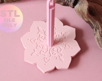 May include: A light pink, flower-shaped stand with a central vertical post. The base features an embossed, symmetrical pattern of swirls and heart shapes. A circular graphic in the upper left corner contains the text STL FILE ONLY.