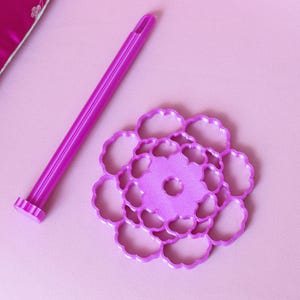 May include: A bright purple plastic tool with a long cylindrical handle and a flat, circular base featuring a cutout floral pattern. The tool is designed for crafting or baking, showcasing a vibrant magenta hue and a detailed, symmetrical design.