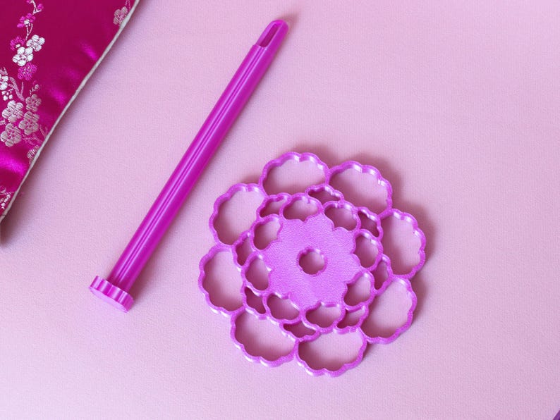 May include: A bright purple plastic tool with a long cylindrical handle and a flat, circular base featuring a cutout floral pattern. The tool is designed for crafting or baking, showcasing a vibrant magenta hue and a detailed, symmetrical design.