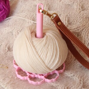 May include: A cream-colored yarn ball sits on a pink, flower-shaped base with a central pink spindle. A brown leather wrist strap with a gold-tone metal lobster clasp is attached to the top of the spindle. Two small gold-tone metal stitch markers are nearby.