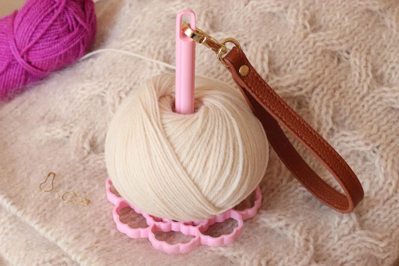 May include: A cream-colored yarn ball sits on a pink, flower-shaped base with a central pink spindle. A brown leather wrist strap with a gold-tone metal lobster clasp is attached to the top of the spindle. Two small gold-tone metal stitch markers are nearby.