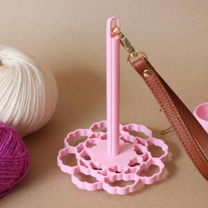 May include: A light pink yarn holder with a scalloped circular base and a vertical spindle. A brown textured wrist strap with a gold-toned metal clasp is attached to the top of the spindle. Two balls of yarn and a small pink container are nearby.