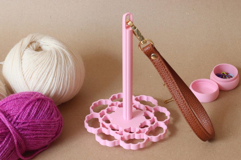 May include: A light pink yarn holder with a scalloped circular base and a vertical spindle. A brown textured wrist strap with a gold-toned metal clasp is attached to the top of the spindle. Two balls of yarn and a small pink container are nearby.