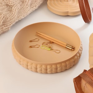 Può includere: Un vassoio rotondo in legno beige con un bordo strutturato e smerlato. All'interno del vassoio ci sono graffette dorate e un paio di bacchette di legno. Un articolo in maglia beige è sullo sfondo, insieme ad altri accessori in legno.