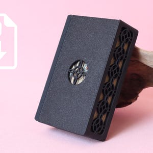 May include: A rectangular black box with a textured surface and a circular cutout in the center. The side panels feature an intricate, repeating geometric pattern of interlocking circles. A white icon with the text STL and a downward arrow is visible.