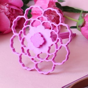 May include: A bright pink plastic flower-shaped cookie cutter featuring a multi-layered petal design with a central textured circular detail. The tool has a handle extending from the back and is constructed with a rigid, open-frame structure.