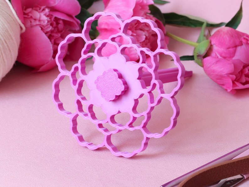 May include: A bright pink plastic flower-shaped cookie cutter featuring a multi-layered petal design with a central textured circular detail. The tool has a handle extending from the back and is constructed with a rigid, open-frame structure.