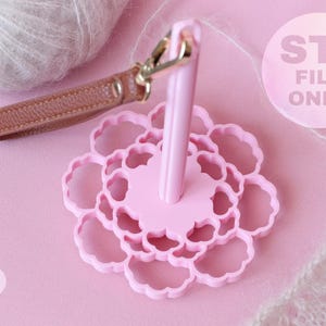 May include: A light pink yarn holder with a flower-shaped base and a central vertical spindle. A brown leather wrist strap with a gold-tone metal lobster clasp is attached to the top of the spindle. The text STL FILE ONLY is visible.