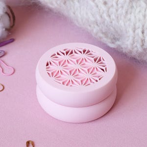 May include: A light pink, round, two-piece container with a geometric design on the top. The container is sitting on a pink surface, with crochet hooks, stitch markers, and yarn in the background.