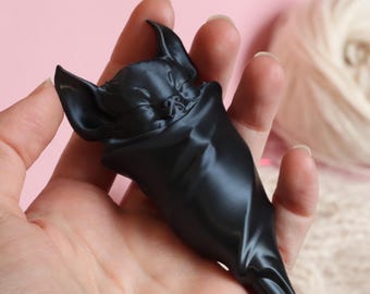 May include: A black bat figurine with large ears and a sleeping expression. The bat is wrapped in a dark, shiny material, creating a sleek, smooth appearance. The figurine is held in a hand against a soft pink background.