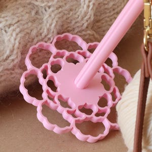 May include: A light pink drop spindle featuring a circular, flower-shaped whorl with scalloped edges and a central shaft. The tool is positioned near a ball of cream-colored yarn and a brown leather strap with a gold-toned metal swivel clasp.