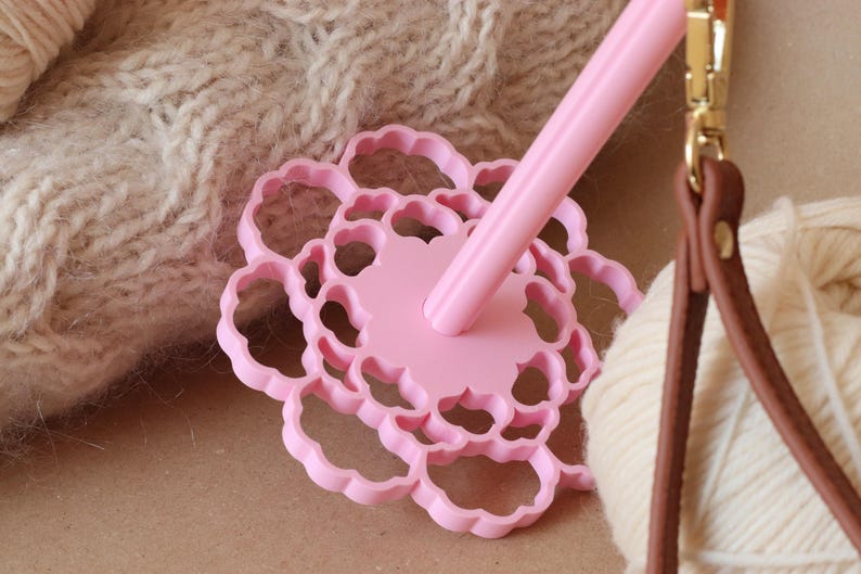 May include: A light pink drop spindle featuring a circular, flower-shaped whorl with scalloped edges and a central shaft. The tool is positioned near a ball of cream-colored yarn and a brown leather strap with a gold-toned metal swivel clasp.