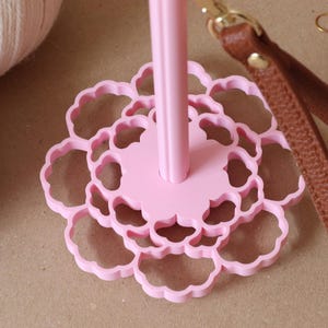 May include: A light pink yarn holder featuring a central vertical spindle attached to a flat, circular base with a scalloped, flower-like cutout pattern. A brown leather strap with a gold-toned metal clasp is attached to the top of the spindle.