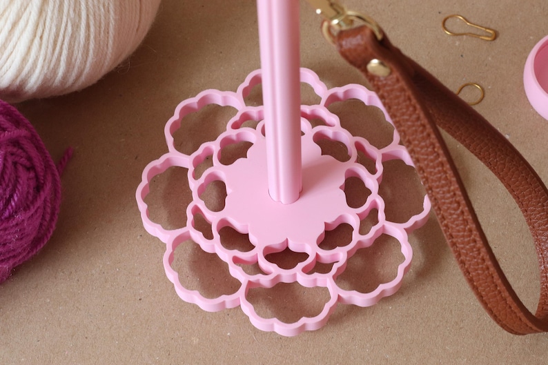 May include: A light pink yarn holder featuring a central vertical spindle attached to a flat, circular base with a scalloped, flower-like cutout pattern. A brown leather strap with a gold-toned metal clasp is attached to the top of the spindle.