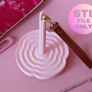 Könnte beinhalten: Ein hellrosa Garnhalter mit einem braunen Lederriemen und einem goldfarbenen Verschluss. Der Halter hat ein blütenförmiges Design. Das Bild enthält auch ein rosa Etui, eine Schere und einen Häkelhaken. Der Text "STL FILE ONLY" ist sichtbar.