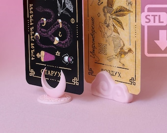 Puede incluir: Dos soportes de tarjetas de color rosa claro sostienen cartas de tarot rectangulares. Un soporte tiene forma de media luna sobre una base circular, mientras que el otro tiene forma de nube estilizada. Las cartas muestran ilustraciones.