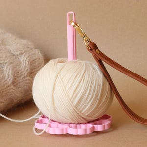 May include: A light pink yarn holder with a vertical spindle and a flower-shaped base. A ball of cream-colored yarn is mounted on the spindle. A brown leather wrist strap with a gold-toned metal lobster clasp is attached to the top of the spindle.