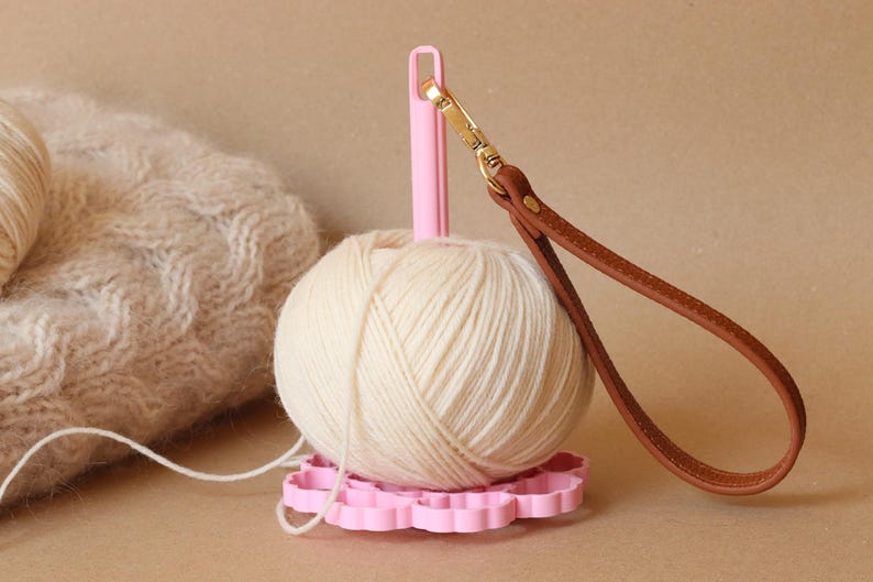 May include: A light pink yarn holder with a vertical spindle and a flower-shaped base. A ball of cream-colored yarn is mounted on the spindle. A brown leather wrist strap with a gold-toned metal lobster clasp is attached to the top of the spindle.