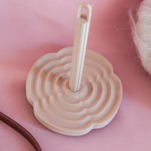 Puede incluir: Un devanador de hilo beige claro con una base en forma de flor y un poste vertical central con un lazo en la parte superior. El devanador está diseñado para sujetar madejas de hilo. El fondo es rosa.