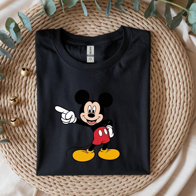 May include: Black t-shirt featuring a colorful graphic of Mickey Mouse pointing. Mickey Mouse is depicted with black ears, a red shorts, yellow shoes, and white gloves. The shirt is folded neatly on a woven surface.