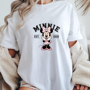 Może przedstawiać: Biała koszulka z grafiką Myszki Minnie. Projekt zawiera słowo "MINNIE" nad wizerunkiem Myszki Minnie w r&oacute;żowej sukience w kropki i kokardą. Tekst "EST. 1928" jest r&oacute;wnież dołączony.