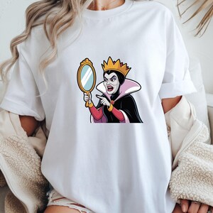 May include: White t-shirt featuring a cartoon image of the Evil Queen from Snow White. The queen is holding a gold-framed mirror and pointing. She wears a crown, black cape, and a pink and white collar.