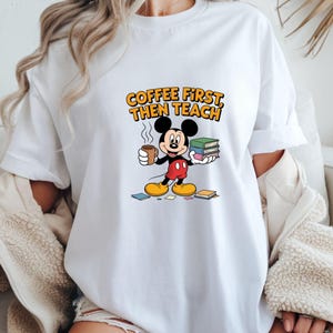 May include: White t-shirt with a cartoon Mickey Mouse design. Mickey is holding a coffee cup and books. The text "COFFEE FIRST THEN TEACH" is printed above the character in orange and yellow.
